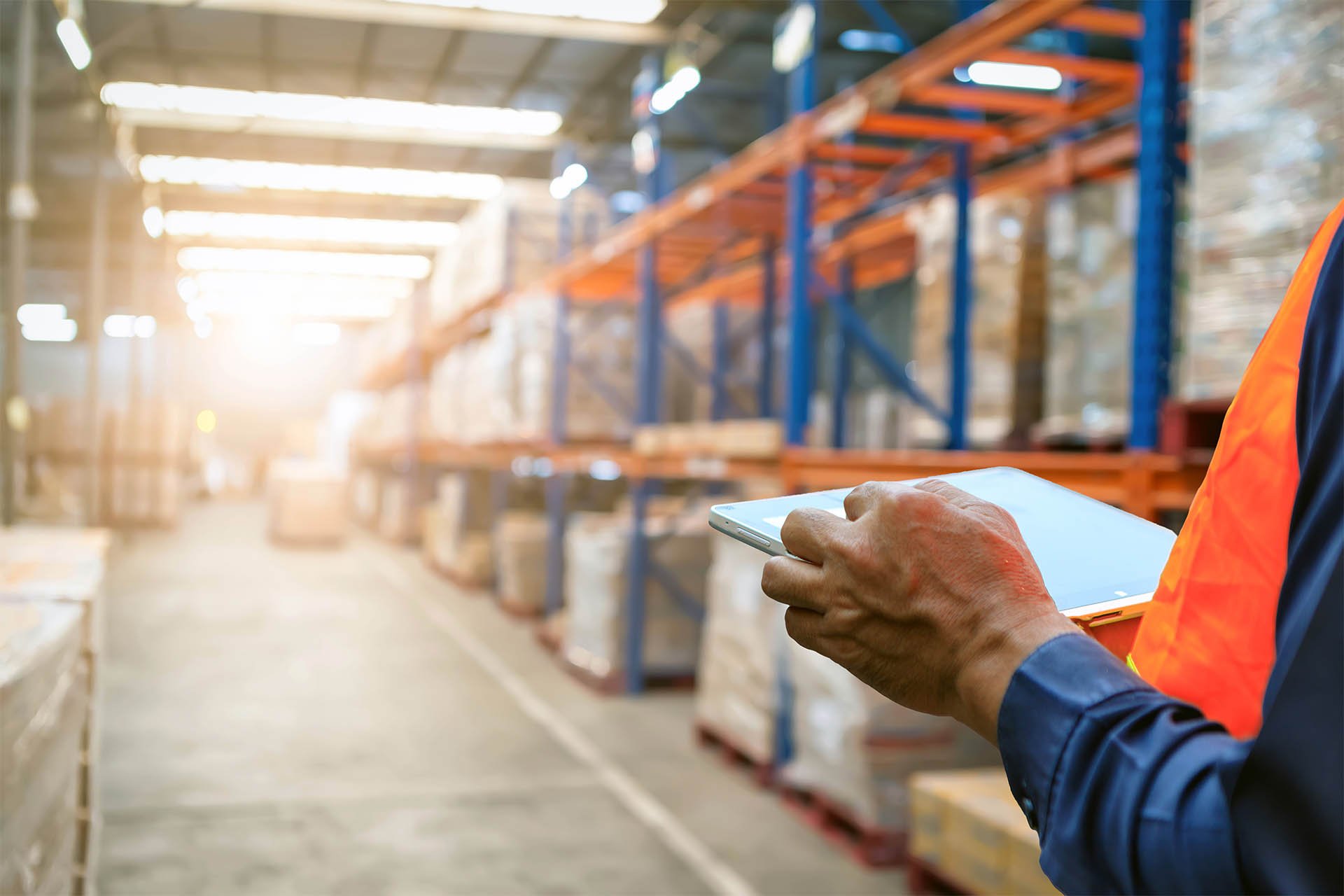 A worker in an orange vest uses a tablet in a well-lit warehouse. Tall shelves are filled with pallets and boxes, creating a sense of organized efficiency.