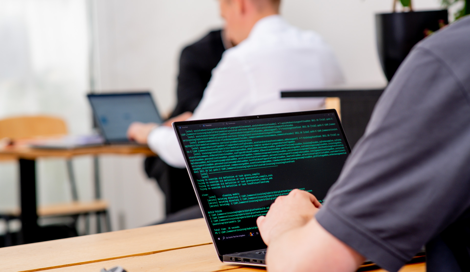A person is typing on a laptop displaying green coding text, with another individual working on a laptop in the background.