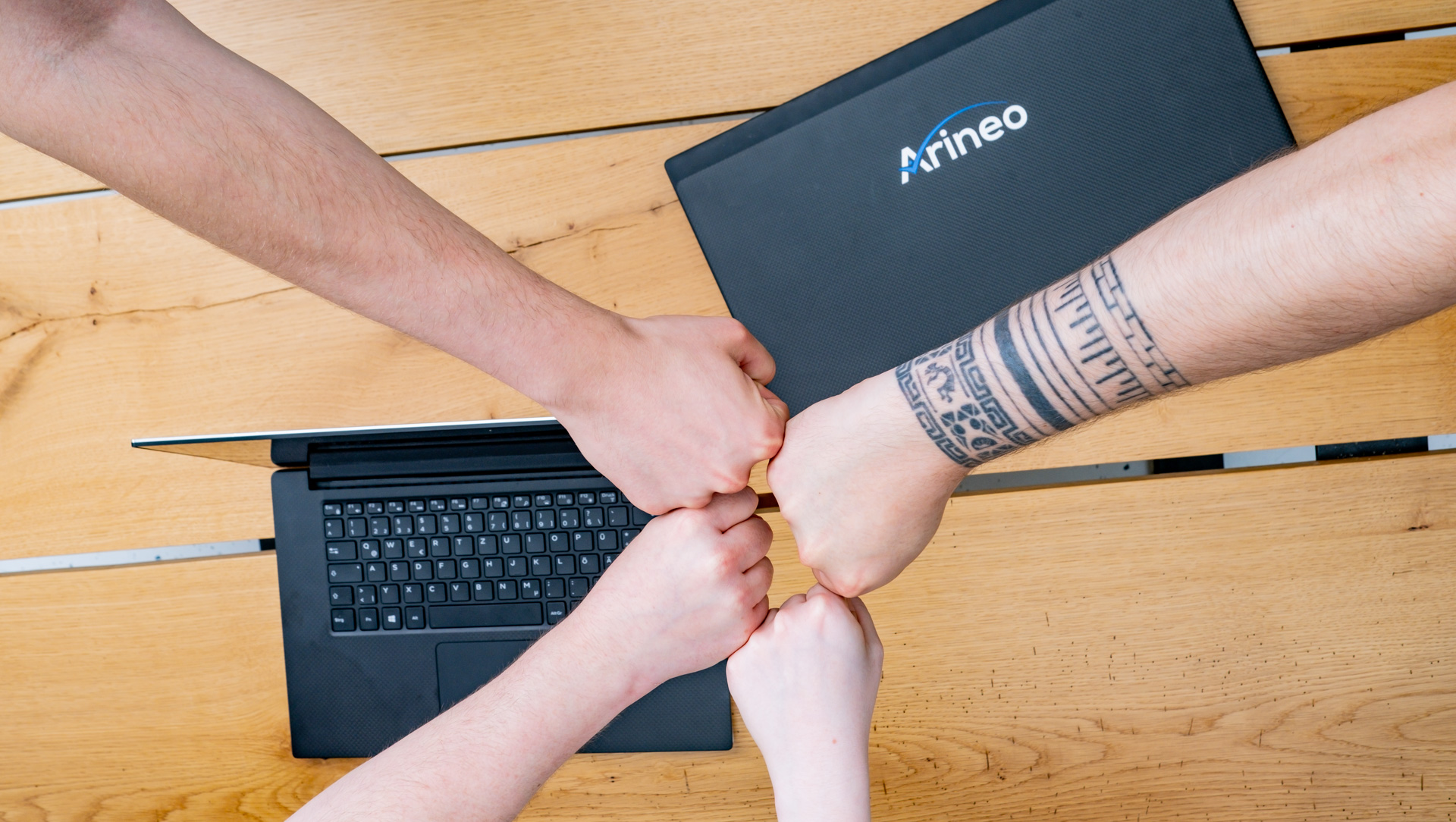Four clasped fists above a laptop on a wooden table, symbolizing teamwork and collaboration. One arm features a distinct tattoo.
