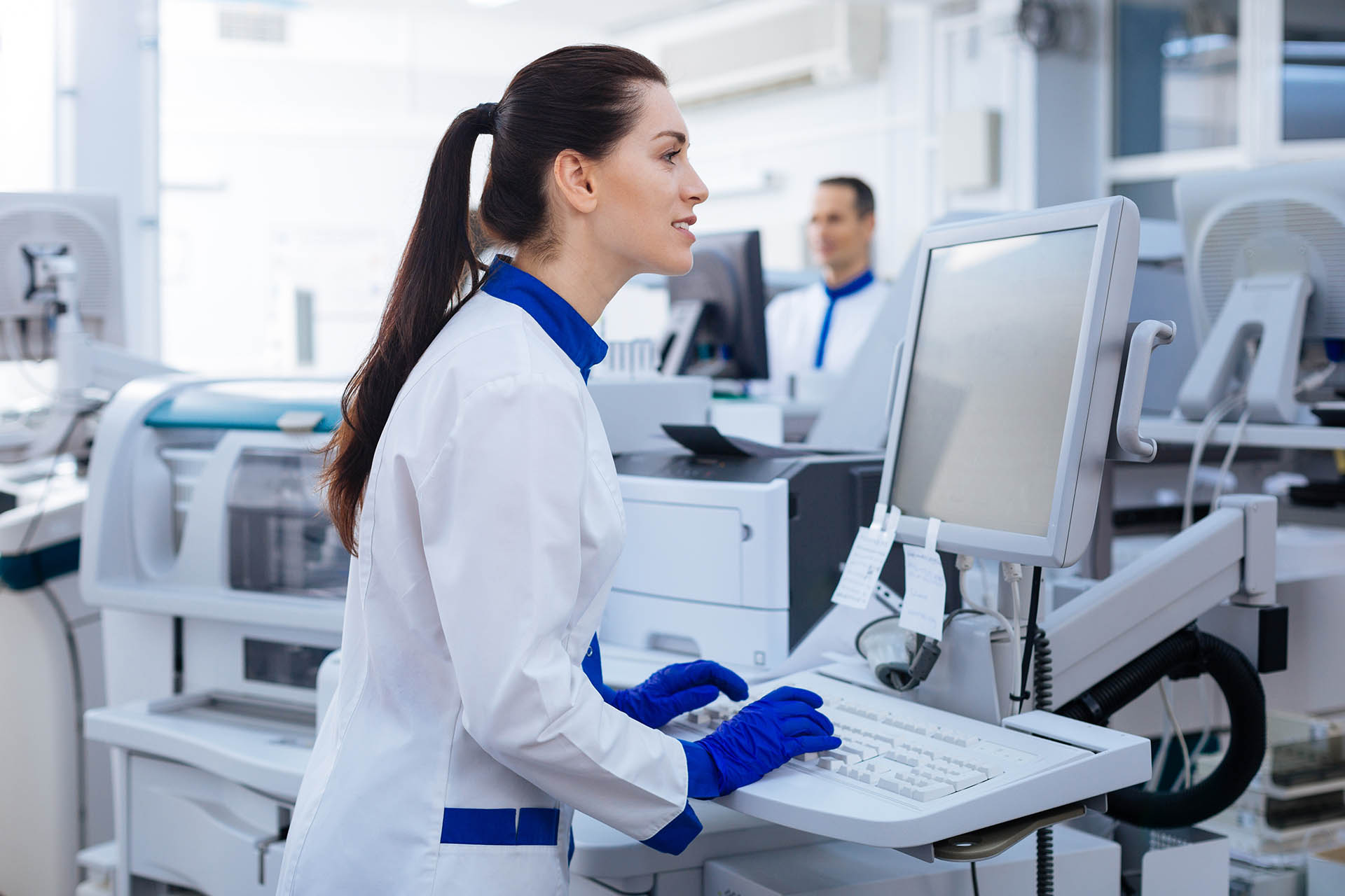 A laboratory technician in a white coat and blue gloves works at a computer, surrounded by medical equipment in a bright workspace.